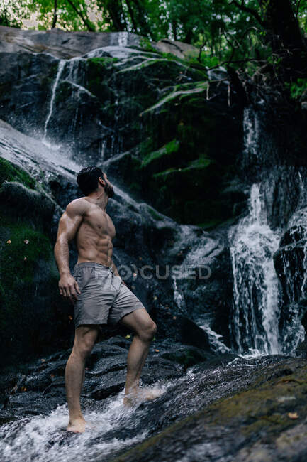 Vista lateral do homem em forma com tronco nu em pé na rocha na água da cachoeira em madeiras — Fotografia de Stock