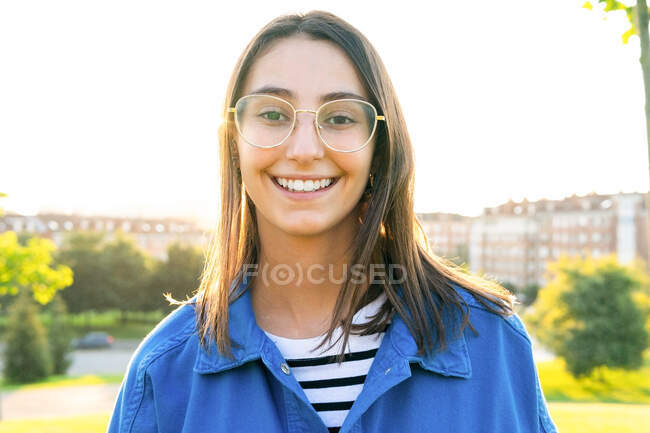 Positive junge Frau in trendiger Kleidung steht auf einem grünen Hügel vor dem Hintergrund der Stadt an einem sonnigen Tag und blickt in die Kamera im Gegenlicht — Stockfoto