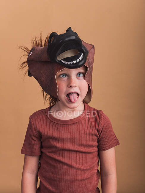 Kind in T-Shirt und Pferdemaske auf dem Kopf blickt in die Kamera auf beigem Hintergrund und streckt die Zunge heraus — Stockfoto