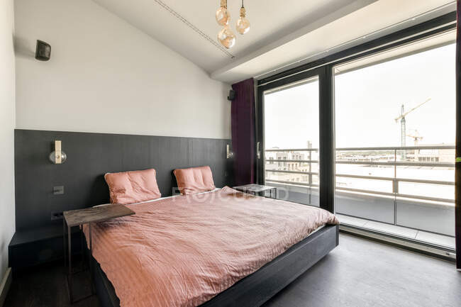 Современный интерьер светлой спальни с большой кроватью, покрытой розовым одеялом, расположенной возле панорамного окна в современной городской квартире в стиле лофт — стоковое фото