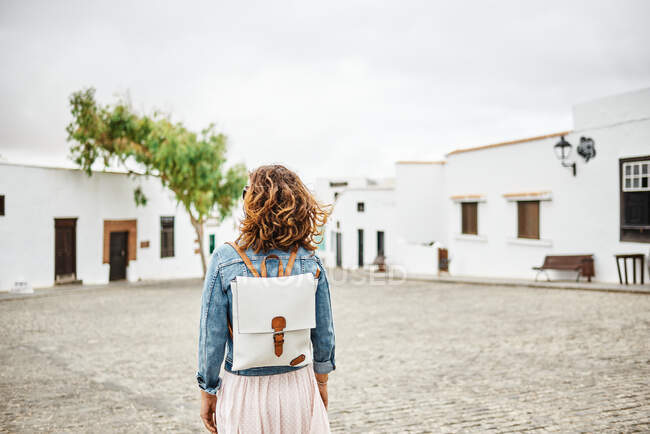 Задний вид анонимной женщины с рюкзаком, идущей по тротуару против белых домов и облачно-серого неба на городской улице в Фуэртевентуре, Испания — стоковое фото