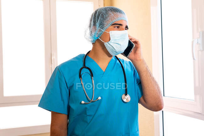 Médico masculino en uniforme y con estetoscopio de pie en el hospital y hablando en el teléfono inteligente - foto de stock