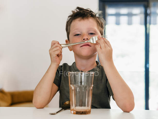Cute preteen child playing with spoon as mustache while sitting at table in kitchen and looking at camera — Stock Photo
