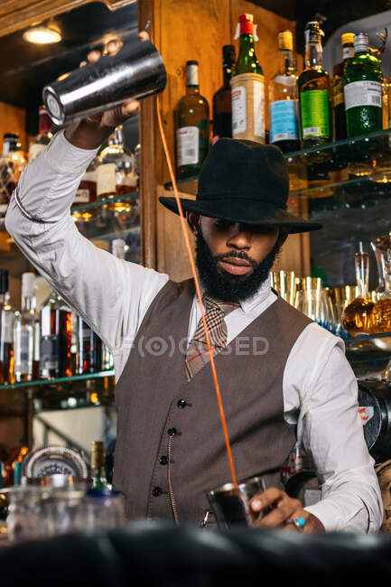 Stylish bearded African American male barkeeper pouring red drink into shaker glass at counter in bar — Stock Photo