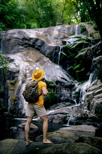 Side view of unrecognizable male hiker standing on boulder and admiring waterfall in forest — Stock Photo