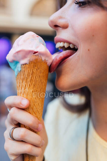 Colheita sorrindo jovem fêmea lambendo saboroso gelato em cone de waffle enquanto olha para longe na cidade no fundo borrado — Fotografia de Stock
