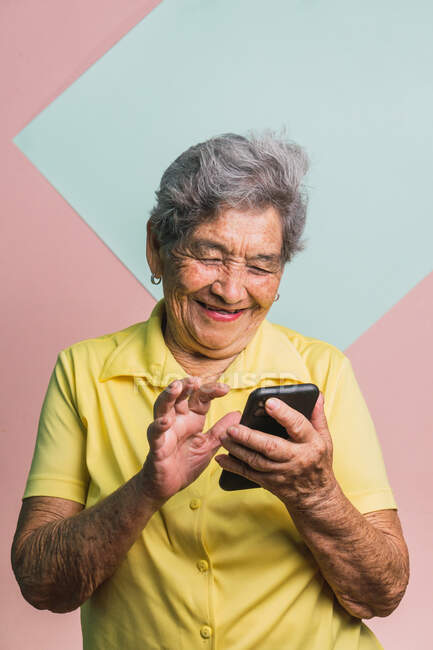 Восхитительная современная пожилая женщина с седыми волосами, просматривающая социальные сети на мобильном телефоне на двух цветном фоне в студии — стоковое фото
