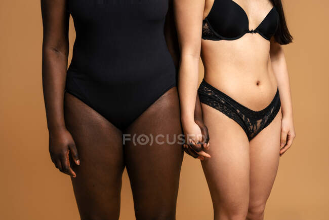 Обрезание неузнаваемых многорасовых женщин в черном нижнем белье держаться за руки, стоя на бежевом фоне в студии для концепции положительного тела — стоковое фото