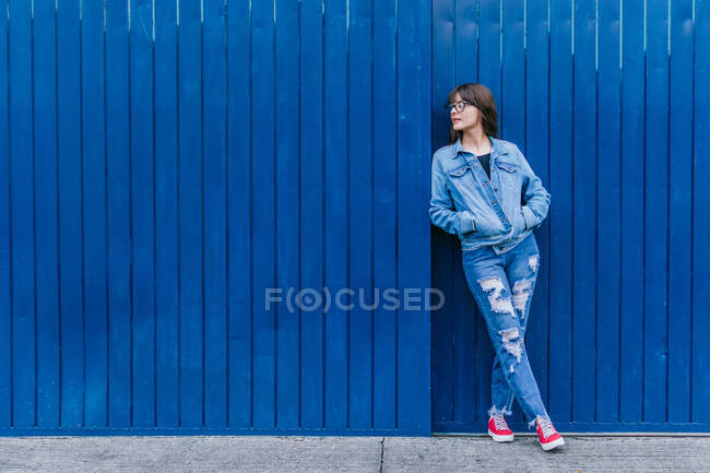 Trendy female hipster wearing denim jacket and jeans leaning on blue wall in city street and looking away — Stock Photo