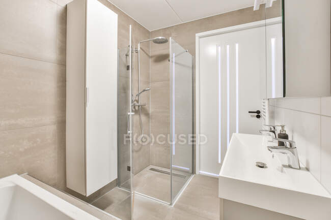 Интерьер домашней ванной комнаты с зеркалом, висящим над двойной раковиной, размещенной рядом с входной дверью и стеклянной душевой кабиной в современной квартире — стоковое фото