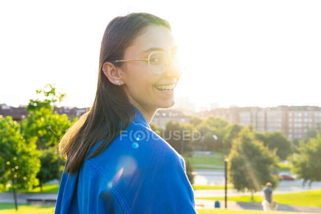 Задний вид женщины в модной одежде стоящей, смотрящей в камеру на зеленом холме на фоне города в солнечный день — стоковое фото