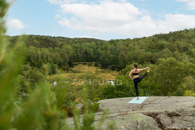 Mulher irreconhecível com a mão na cintura fazendo estendido mão para Big Toe pose na pedra no campo exuberante no verão — Fotografia de Stock