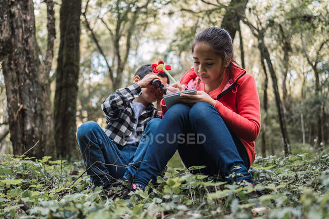 Етнічна дівчина пише в блокноті проти брата, дивлячись через бінокль, сидячи на землі в літньому лісі — стокове фото