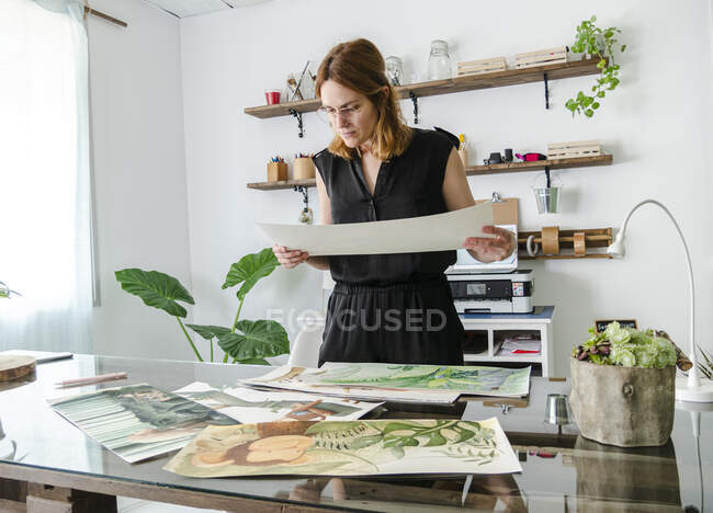 Creative female designer examining various paintings placed on table and working on project — Stock Photo
