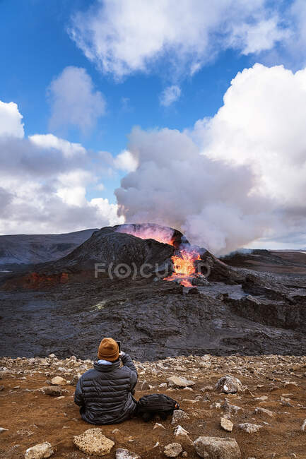 Back view of faceless tourist taking photo of active Fagradalsfjall on cellphone while sitting on mount under cloudy blue sky in Iceland — Stock Photo