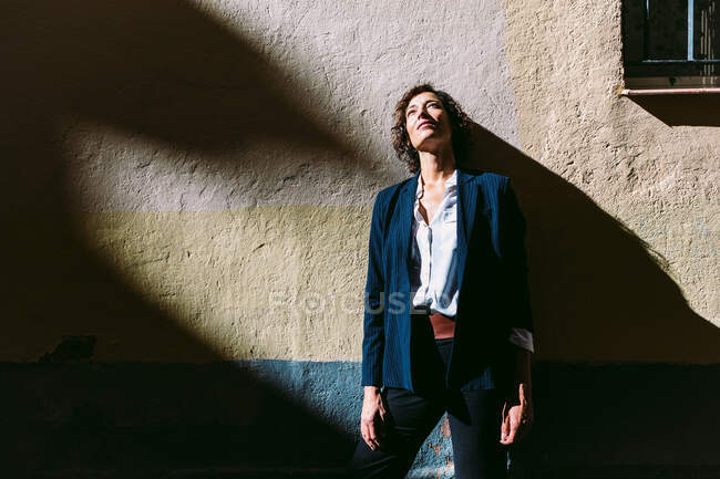 Nachdenkliche Frau in eleganter Kleidung steht im Sonnenlicht und wirft Schatten auf schäbige Wand und schaut weg — Stockfoto