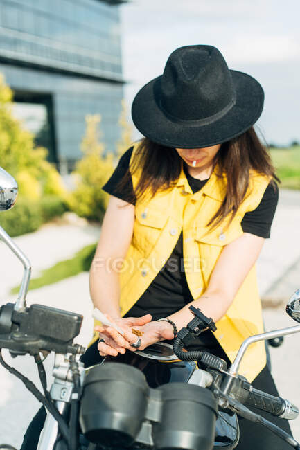 Anonymous female motorcyclist in hat leaning on modern motorbike parked on road in city on sunny day — Stock Photo