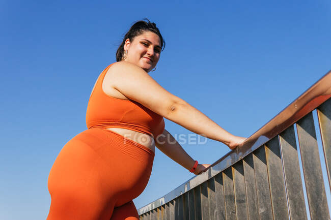 Von unten betrachtet kontemplative ethnische Sportlerin mit kurvigem Körper die Kamera, während sie sich am Zaun unter blauem Himmel lehnt — Stockfoto