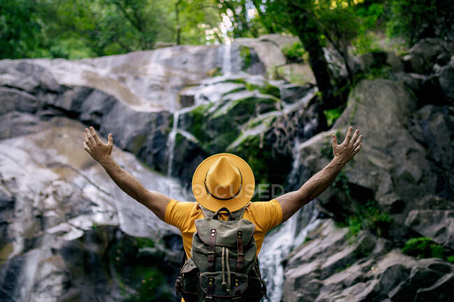 Обратный вид на неузнаваемого туриста-мужчину, стоящего на валуне и любующегося водопадом в лесу с распростертыми объятиями — стоковое фото