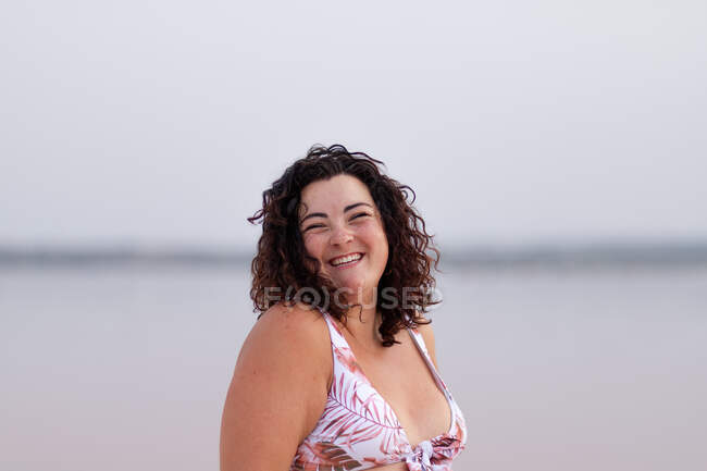 Deliciosa mujer con curvas en bikini de pie cerca del agua de estanque rosa en verano y mirando a la cámara - foto de stock