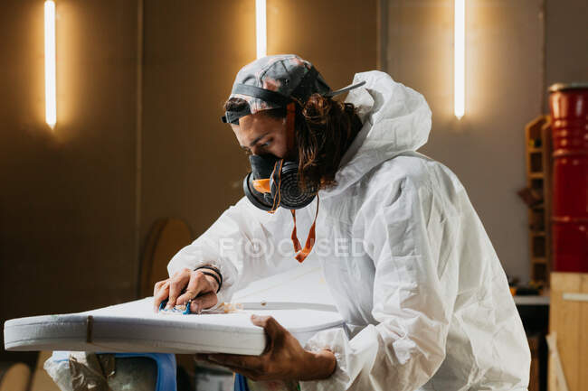 Seitenansicht eines männlichen Shaper in Sicherheitskostüm und Atemschutzmaske poliert Oberfläche des Surfbretts mit Handhobel in der Werkstatt — Stockfoto