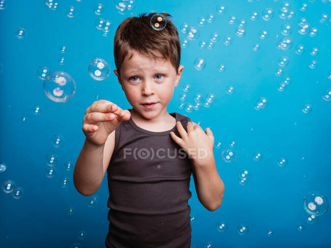 Étonné préadolescent garçon montrant geste touchant avec index dans l'air en studio avec bulles de savon volant sur fond bleu — Photo de stock