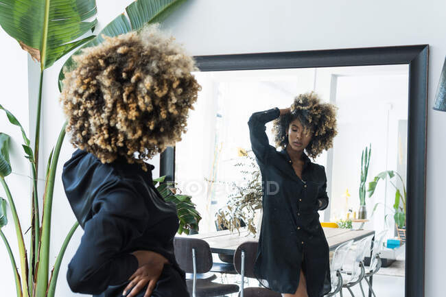 Visão traseira da mulher afro-americana magra em vestido da moda e com cabelo encaracolado refletindo no espelho enquanto está em pé no quarto em casa — Fotografia de Stock