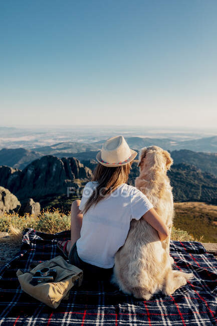Back view of anonymous female traveler embracing Golden Retriever dog and looking at mountains while sitting on checkered blanket in highlands. — Stock Photo