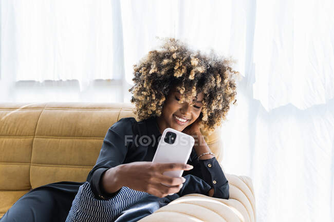 Позитивная афроамериканка с афро-прической, которая делает автопортрет на мобильном телефоне, сидя дома на диване и развлекаясь — стоковое фото