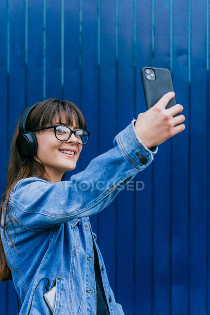 Vista lateral o hipster feminino feliz em fones de ouvido tomando auto tiro no celular no fundo da parede azul na rua — Fotografia de Stock