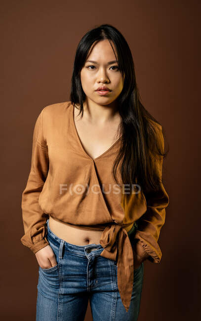 Передній погляд азіатської жінки в модний одяг дивлячись на камеру на коричневому тлі в студії — стокове фото