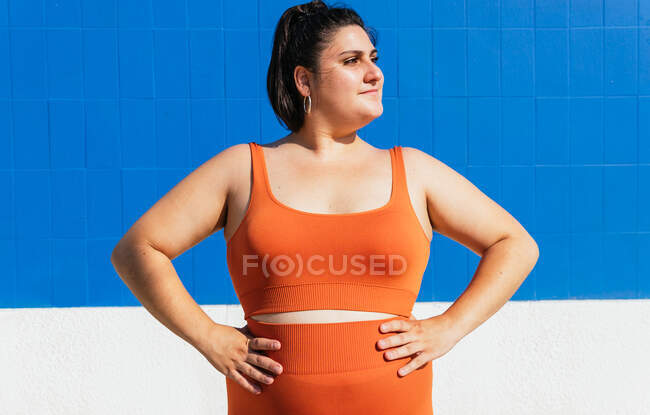 Atleta di etnia femminile sovrappeso in usura attiva guardando lontano contro la parete piastrellata blu nella giornata di sole — Foto stock