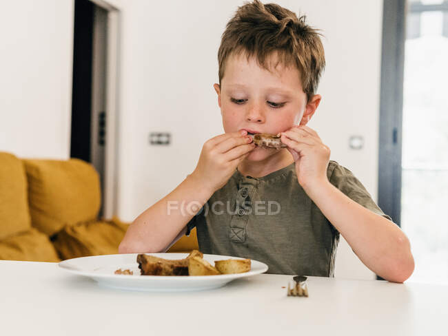 Adorable kid eating appetizing pork ribs during lunch at home and looking down — Stock Photo