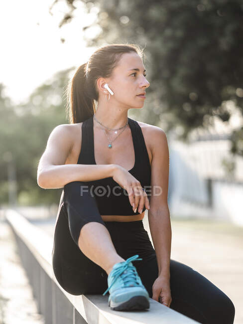 Молодая спортсменка слушает музыку в наушниках TWS и смотрит в сторону, сидя на барьере во время перерыва в фитнес-тренировке — стоковое фото