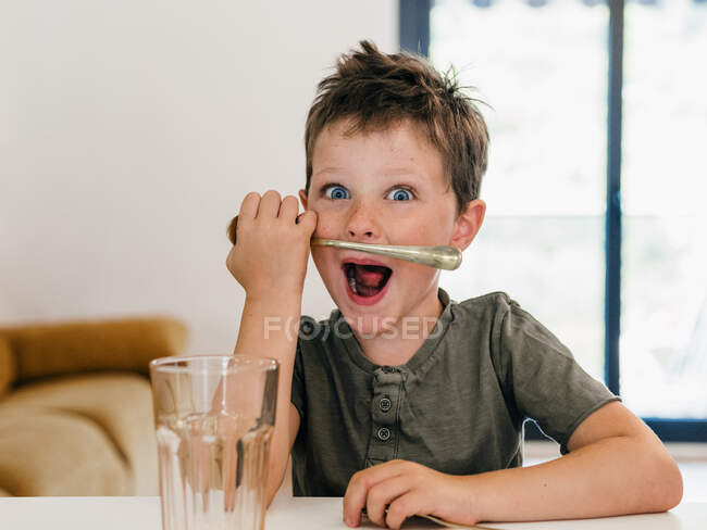 Lindo niño preadolescente jugando con cuchara como bigote mientras está sentado en la mesa en la cocina y mirando a la cámara - foto de stock
