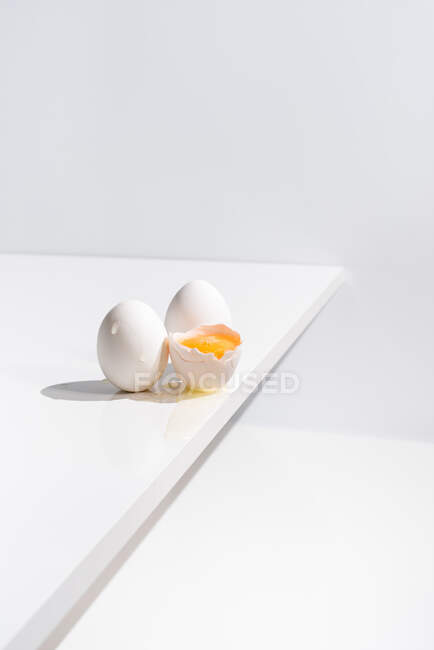 Високий кут цілого і розбитого яйця з жовтком в оболонці, розміщені на краю столу на білому тлі в студії — стокове фото