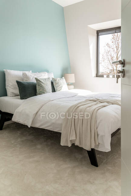Интерьер современной спальни с кроватью с мягкими подушками помещен рядом с окном в квартире в минимальном стиле — стоковое фото