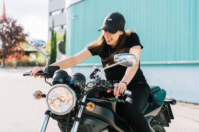 Motociclista feminina positiva sentada em moto estacionada na rua da cidade no dia ensolarado e olhando para longe — Fotografia de Stock