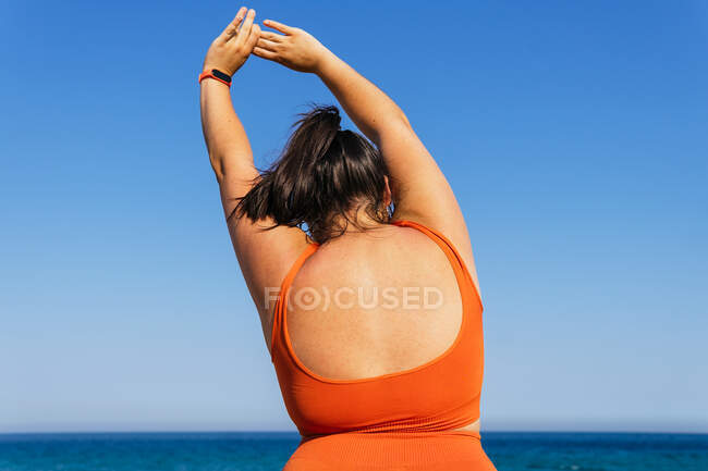 Back view of anonymous plump female athlete in sportswear exercising with raised arms against ocean under blue sky — Stock Photo