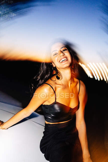 Attraktive, glückliche junge, langhaarige hispanische Brünette in schwarzem Top mit nackten Schultern, die in die Kamera schaut, die nachts in einem Auto mit Lichtreflexion sitzt — Stockfoto