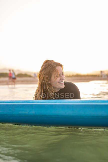 Mujer surfista acostada sobre tabla de SUP y flotando en aguas tranquilas de mar en un día soleado mirando hacia otro lado durante la puesta del sol - foto de stock