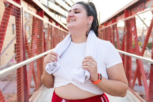Cheerful plus size ethnic female athlete in sportswear with towel looking away during training on bridge in town — Stock Photo