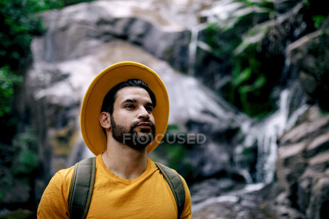 Vista laterale dell'escursionista maschio in piedi sul masso e accanto alla cascata e guardando lontano nella foresta — Foto stock