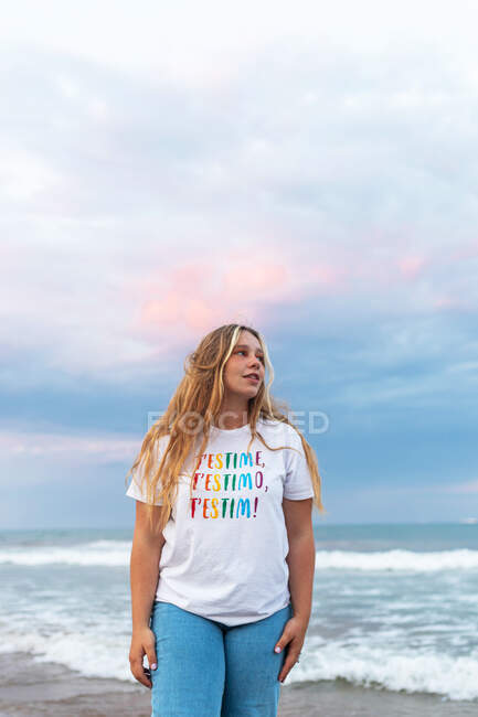 Von unten positive junge Frau in stylischem Outfit, die an einem Sommerabend am Meer steht — Stockfoto