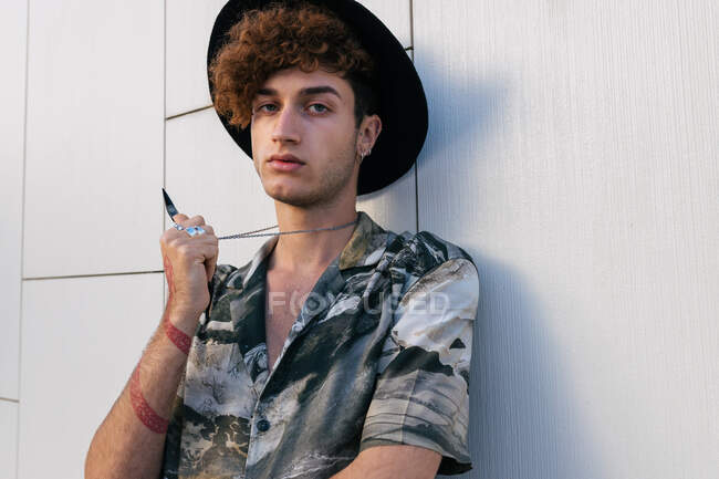 Молодой тщеславный мужчина в стильной одежде в шляпе, стоящий на черепичной стене и смотрящий в камеру — стоковое фото