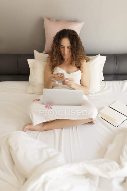 Von oben sitzt Geschäftsfrau mit lockigem Haar im Bett und arbeitet mit Laptop und Smartphone — Stockfoto