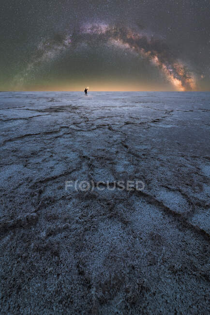 Distant silhouette of explorer standing and holding a flashlight in dry salt lagoon on background of starry sky with glowing Milky Way at night — Stock Photo