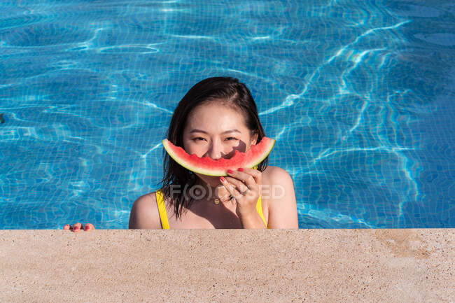 Fröhliche ethnische Frau im Pool, die ihr Gesicht mit einer Scheibe Wassermelone bedeckt, während sie an einem sonnigen Sommertag in die Kamera blickt — Stockfoto