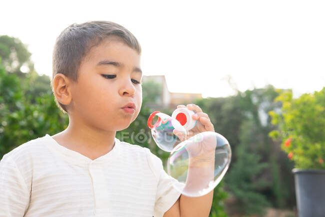 Милый маленький мальчик в белой футболке, надувающий мыльные пузыри, стоя в зеленом саду в летний день — стоковое фото