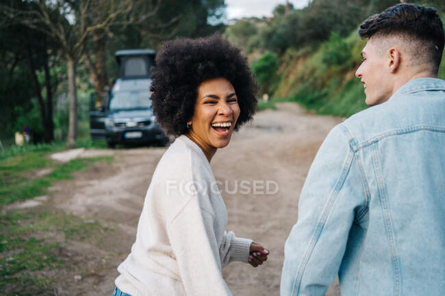 Lächelnde Afroamerikanerin hält Händchen von beschnittenem Freund beim Spazierengehen und schaut gegen Wohnmobil weg — Stockfoto